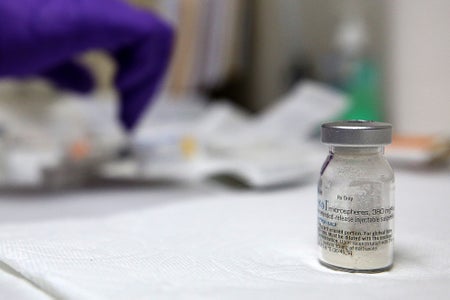 Close-up of a glass vial containing Vivitrol, a prescription drug, standing on a table. A nurse's gloved hand is seen in the background preparing injections