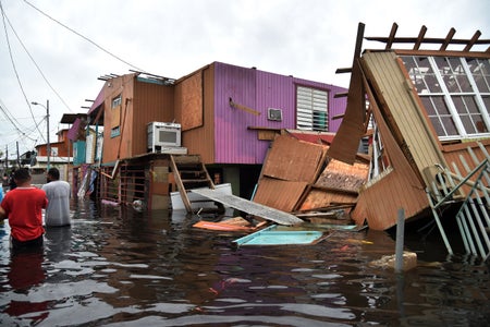 Two men in red and grey shirt wading in water through a street with wood coming off damaged houses.