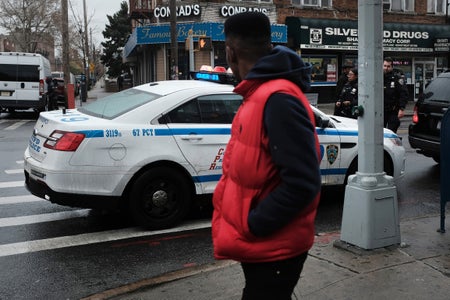 A young black man wearing a red vest looks away from the camera towards a police car parked on a street corner as he walks by in the in the Brownsville neighborhood of Brooklyn on November 18, 2019 in New York City