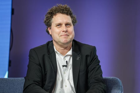 Peter Beck sitting in a blue chair on stage at a summit with a purple background