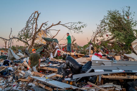 A man in a green shirt walking through the destruction of a home with damaged trees at sunset.