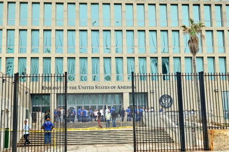 A view of personnel in front of the entrance to the U.S. Embassy in Havana, Cuba, looking through an open black metal bar security gate, chain link fence, and caution tape